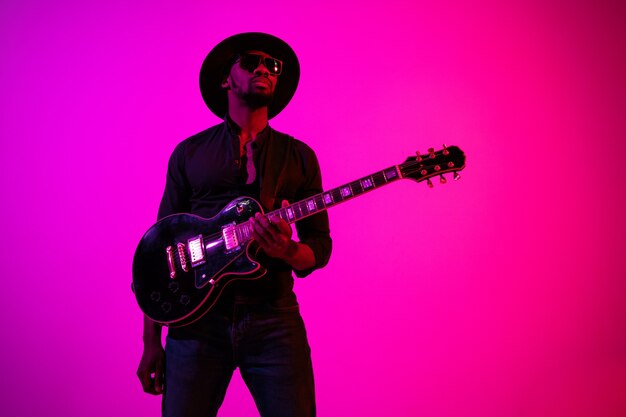 Joven músico afroamericano tocando la guitarra como una estrella de rock sobre fondo degradado de color rosa púrpura en luz de neón.