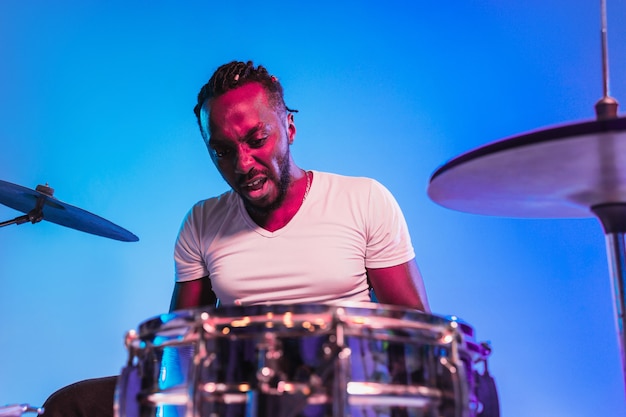 Joven músico de jazz afroamericano o baterista tocando la batería sobre fondo azul de estudio en luces de neón de moda. Concepto de música, afición, inspiración. Retrato colorido del artista alegre.