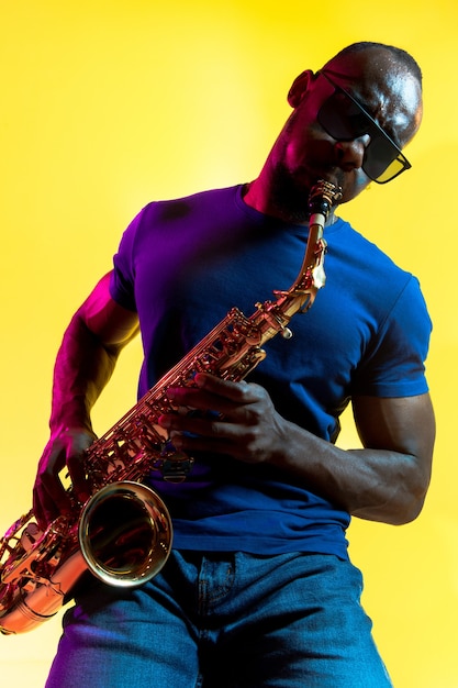 Joven músico de jazz afroamericano tocando el saxofón sobre fondo amarillo en luz de neón de moda.