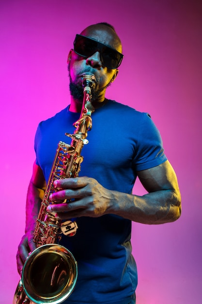 Foto gratuita joven músico de jazz afroamericano tocando el saxofón sobre fondo rosa estudio en luz de neón de moda. concepto de música, afición. chico alegre improvisando. retrato colorido del artista.