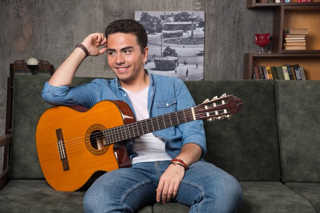 Joven músico sosteniendo una hermosa guitarra y sentado en el sofá. Foto de alta calidad