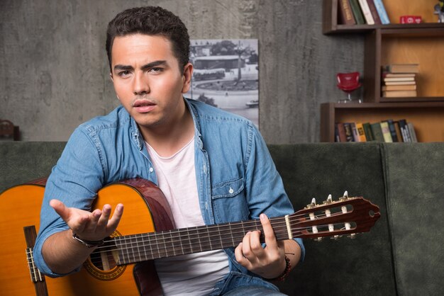 Joven músico sosteniendo una hermosa guitarra y sentado en el sofá. Foto de alta calidad