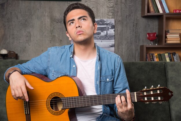 Joven músico sosteniendo una hermosa guitarra y sentado en el sofá. Foto de alta calidad