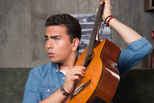 Joven músico sosteniendo una hermosa guitarra y sentado en el sofá. Foto de alta calidad