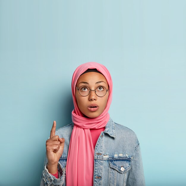 La joven musulmana sorprendida apunta hacia arriba con un dedo índice, enfocado hacia arriba, tiene expresión facial preocupada, cabeza velada