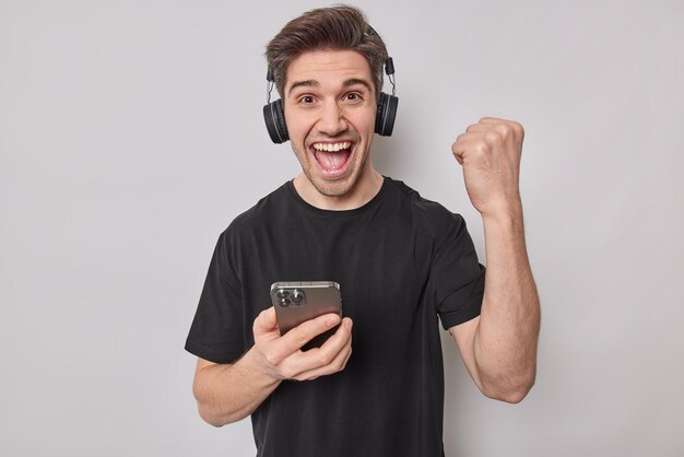 El joven muy contento aprieta el puño con el triunfo contento de ganar el juego en línea usa tecnologías modernas exclama de alegría sostiene el teléfono móvil usa auriculares estéreo en las orejas vestido con una camiseta negra