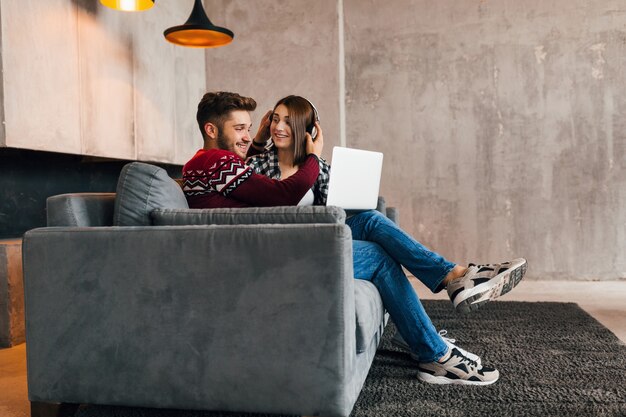 Joven muy feliz sonriente hombre y mujer sentados en casa en invierno, mirando en la computadora portátil, escuchando auriculares, estudiantes que estudian en línea, pareja en el tiempo libre juntos