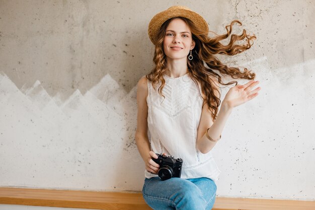 Joven muy sonriente feliz mujer vistiendo camisa blanca sentada contra la pared con sombrero de paja