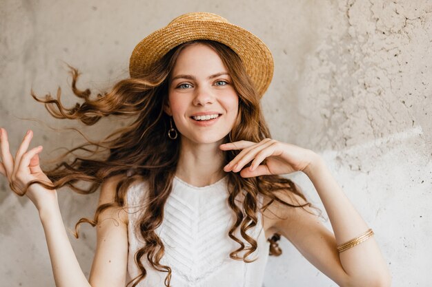 Joven muy sonriente feliz mujer vistiendo camisa blanca sentada contra la pared con sombrero de paja