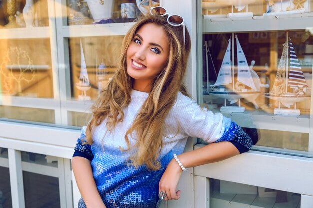 Joven muy sonriente posando cerca de la tienda de souvenirs con barcos y productos de marineros, vistiendo un suéter ombre, maquillaje natural y peinado rizado esponjoso. Disfrute de un buen día y de compras.