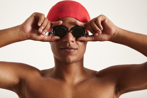 Joven nadador afroamericano serio en gorra roja poniéndose gafas de natación negras sobre blanco