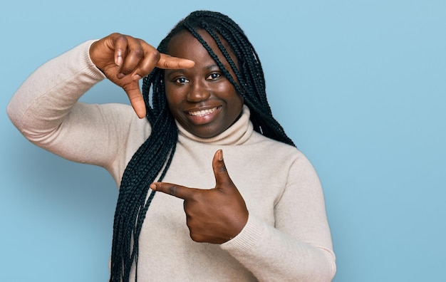 Foto gratuita joven negra con trenzas usando un suéter casual de invierno sonriendo haciendo marco con manos y dedos con cara feliz. concepto de creatividad y fotografía.