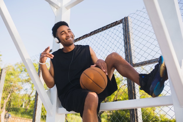 Joven negro haciendo deporte, jugando baloncesto, estilo de vida activo, mañana de verano, sonriendo feliz divirtiéndose escuchando música en auriculares