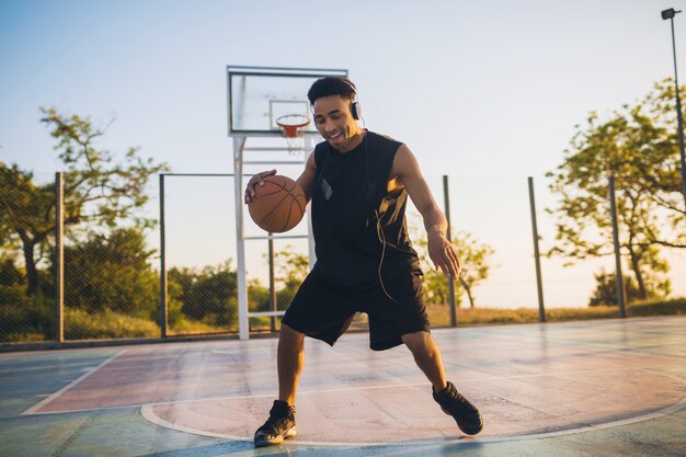 Joven negro sonriente feliz haciendo deporte, jugando baloncesto al amanecer, escuchando música en auriculares, estilo de vida activo, mañana de verano