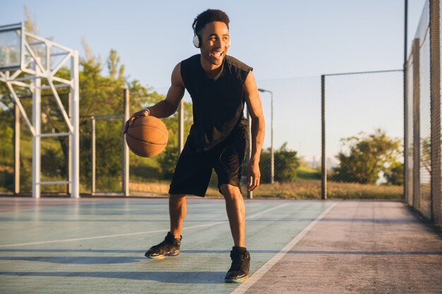 Joven negro sonriente feliz haciendo deporte, jugando baloncesto al amanecer, escuchando música en auriculares, estilo de vida activo, mañana de verano