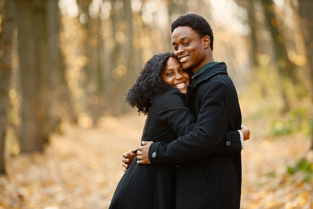 Joven negro y su novia abrazándose. Pareja romántica caminando en el parque de otoño. Hombre y mujer con abrigos negros.