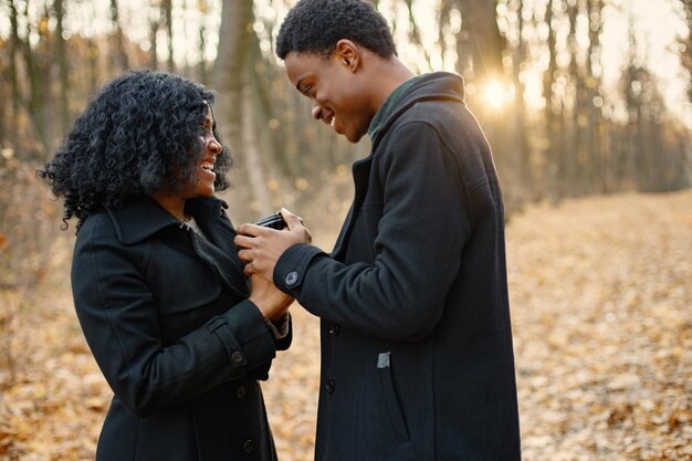 Joven negro y su novia sosteniendo en las manos una taza de café. Pareja romántica caminando en el parque de otoño. Hombre y mujer con abrigos negros.