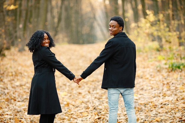 Joven negro y su novia tomados de la mano. Pareja romántica caminando en el parque de otoño al atardecer. Hombre y mujer con abrigos negros.