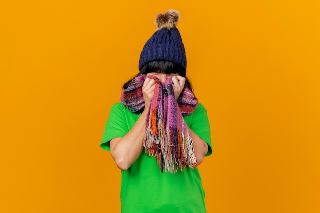 Joven niña caucásica enferma con gorro de invierno y bufanda mirando a cámara sosteniendo bufanda en la cara aislada sobre fondo naranja con espacio de copia