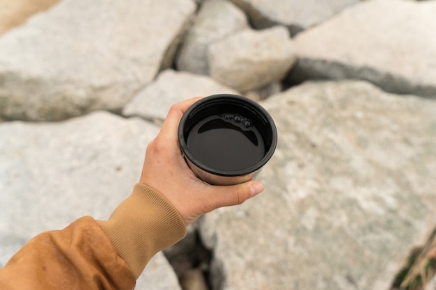 Joven nómada o mujer en busca de aventuras con chaqueta de cuero marrón sostiene una taza de café o té negro americano en una taza de camper o una taza de camping