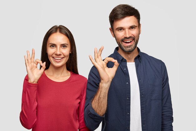 La joven novia y el novio felices muestran el signo de ok con las manos, expresan un símbolo excelente, demuestran su aprobación o acuerdo, tienen expresiones alegres, están de pie en el interior contra la pared blanca