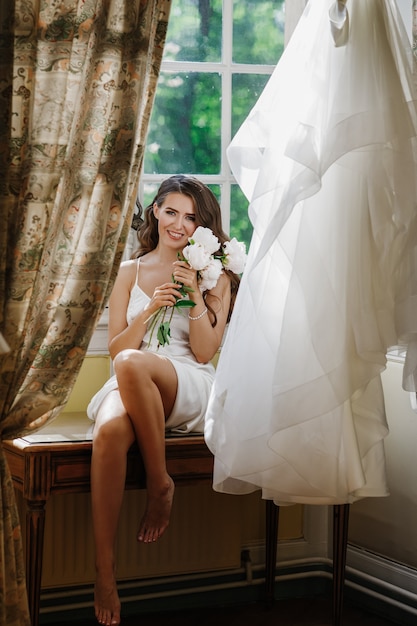 Foto gratuita joven novia tiene un bouuqet de peonías emplazamiento en el alféizar de la ventana en la brillante mañana