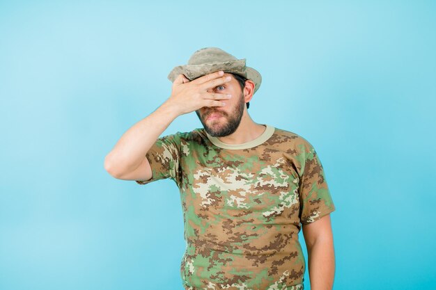 El joven oficial divertido está mirando a través de los dedos en el fondo azul