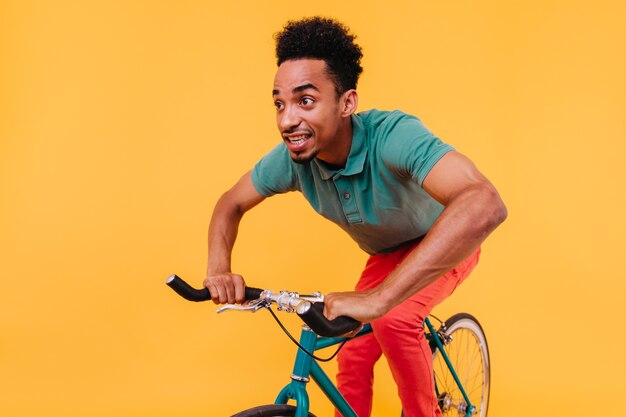 Joven de ojos oscuros en camiseta verde montando en bicicleta. Chico africano feliz en traje casual sentado en bicicleta.