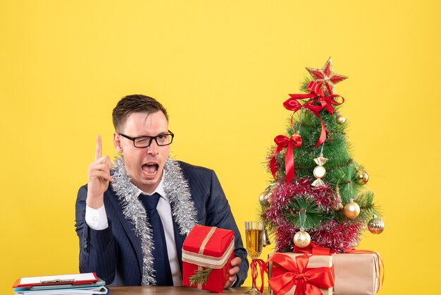 Joven con ojos parpadeados sosteniendo un regalo de Navidad sentado en la mesa cerca del árbol de Navidad y presenta en amarillo