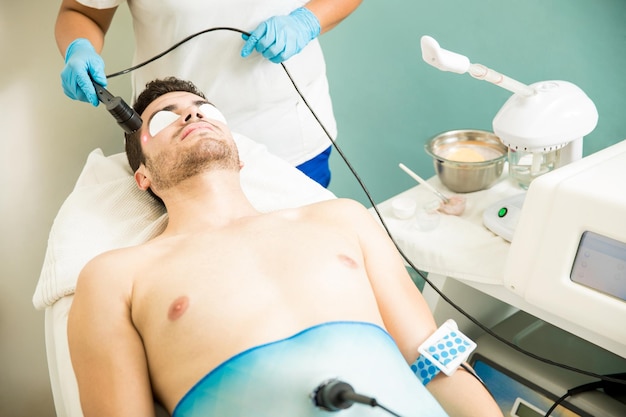 Joven con los ojos tapados recibiendo radiofrecuencia y terapia antienvejecimiento en un spa de salud