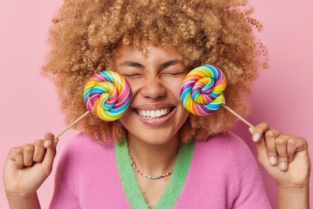 Una joven optimista de cabello rizado mantiene dos piruletas cerca de la cara tiene un diente dulce recuerda el sabor de la infancia disfruta comiendo poses de comida trampa contra un fondo rosa Azúcar nocivo para la salud