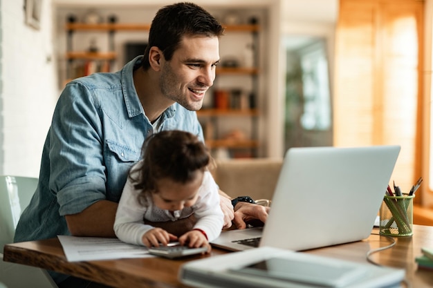 Joven padre feliz leyendo correo electrónico en una computadora mientras está con su hija en casa