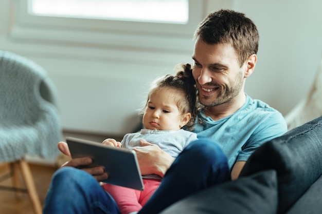 Joven padre feliz usando tableta digital con su pequeña hija mientras se relaja en la sala de estar.