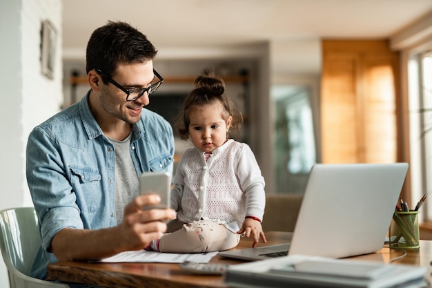 Joven padre trabajador usando teléfono móvil y comunicándose con su pequeña hija en casa