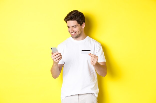 Joven pagando en línea, inserte el número de tarjeta de crédito en el teléfono móvil, comprando en internet, de pie sobre fondo amarillo