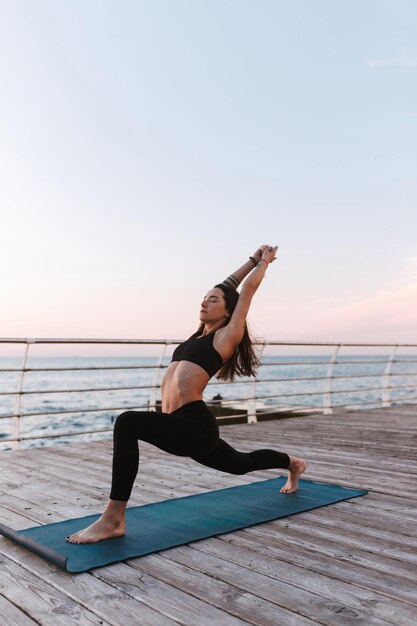 Joven pansiva de pie y entrenando poses de yoga junto al mar. Hermosa mujer en top deportivo negro y polainas practicando yoga con vista al mar en el fondo