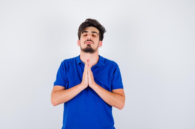 Joven parado en pose de oración en camiseta azul y mirando serio. vista frontal.