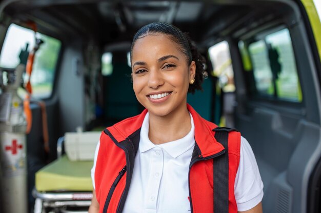 Una joven paramédica parada en la parte trasera de una ambulancia junto a las puertas abiertas Ella está mirando a la cámara con una expresión de confianza sonriendo llevando una bolsa de trauma médico en su hombro