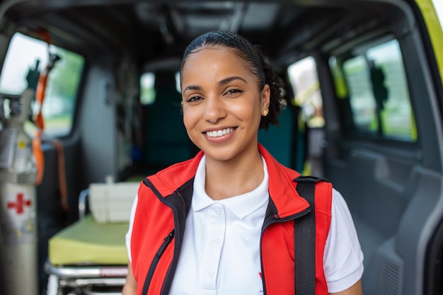 Foto gratuita una joven paramédica parada en la parte trasera de una ambulancia junto a las puertas abiertas ella está mirando a la cámara con una expresión de confianza sonriendo llevando una bolsa de trauma médico en su hombro