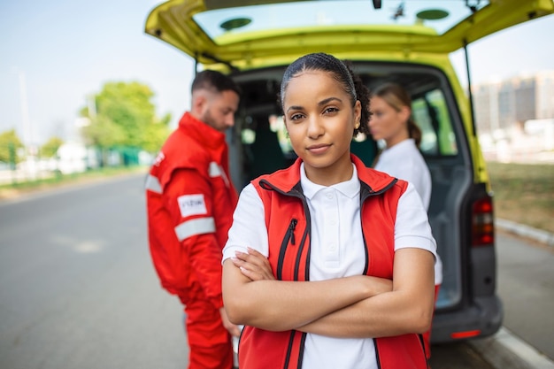 Foto gratuita joven paramédico afroamericano de pie detrás de los paramédicos de la ambulancia junto a la ambulancia dos paramédicos sacando la camilla de la ambulancia