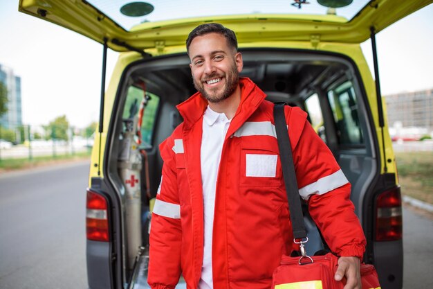 Un joven paramédico parado en la parte trasera de una ambulancia junto a las puertas abiertas Está mirando a la cámara con una expresión de confianza sonriendo llevando una bolsa de trauma médico en el hombro