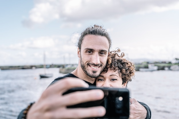 Joven pareja abrazada por el hombro haciendo una selfie con el puerto y el mar desenfocado