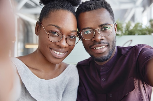 Joven pareja africana hace selfie, se para cerca el uno del otro, expresa emociones positivas, usa gafas.