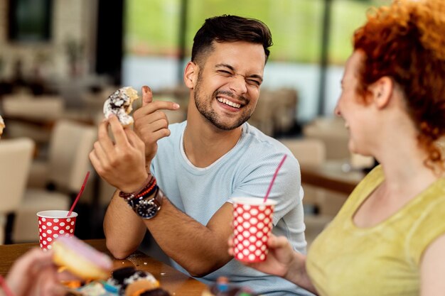 Joven pareja alegre comiendo donuts y divirtiéndose en un café