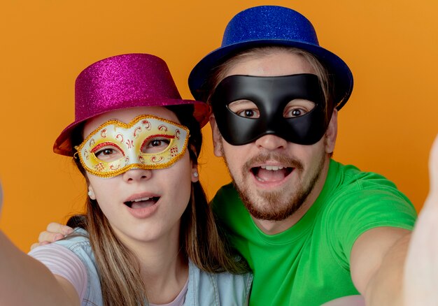 Joven pareja alegre con sombreros rosados y azules puestos en máscaras de ojos de mascarada finge sostener y parece aislado en la pared naranja