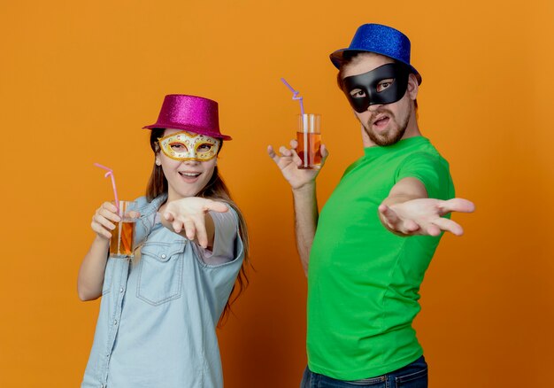 Joven pareja alegre con sombreros rosados y azules se puso máscaras de ojos de mascarada sosteniendo un vaso de jugo extiende las manos aisladas en la pared naranja