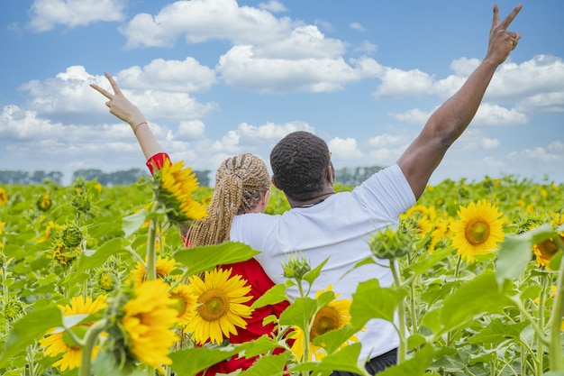 Joven pareja amorosa de pie en medio de un campo de girasoles mostrando un signo de victoria