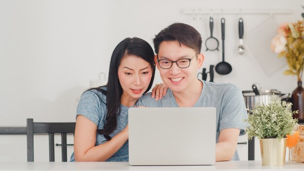 Joven pareja asiática disfruta de compras en línea en la computadora portátil en casa. Estilo de vida joven esposo y esposa felices compran comercio electrónico después de desayunar en la cocina moderna de la casa por la mañana.