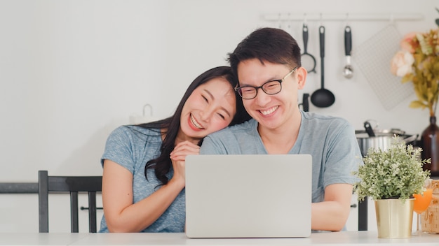 Joven pareja asiática disfruta de compras en línea en la computadora portátil en casa. Estilo de vida joven esposo y esposa felices compran comercio electrónico después de desayunar en la cocina moderna de la casa por la mañana.