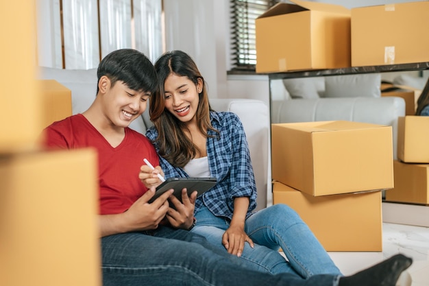 Joven pareja asiática sentada frente a un sofá en el piso y usa una tableta mientras cuenta cajas después de la reubicación en su nuevo hogar nuevo concepto de mudanza